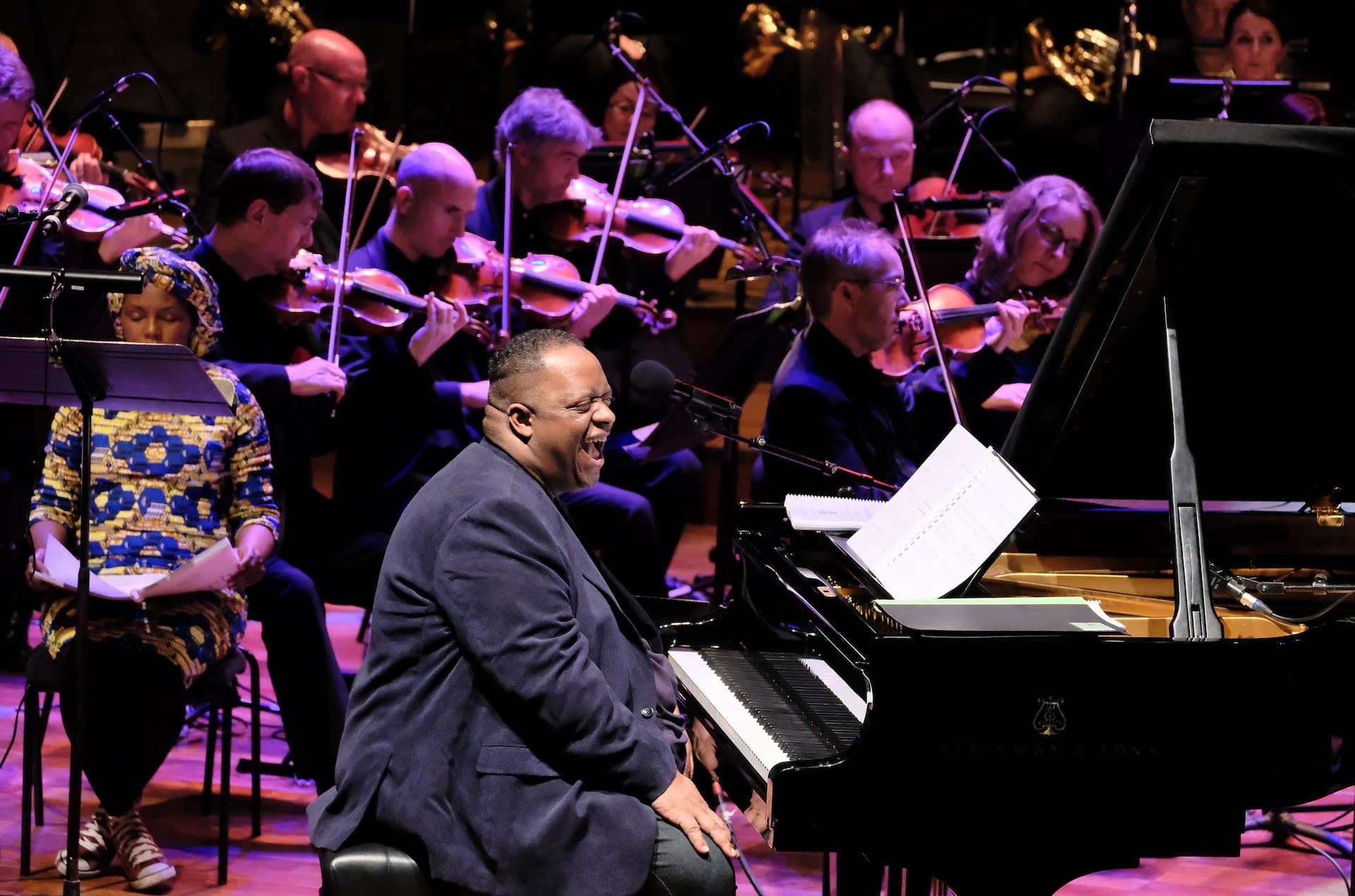 Julian Joseph sits at a piano with an orchestra behind him.