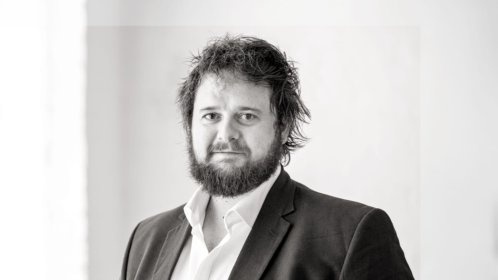 Tenor Allan Clayton, a man with short dark hair and a beard, wearing a dark suit, formal portrait.