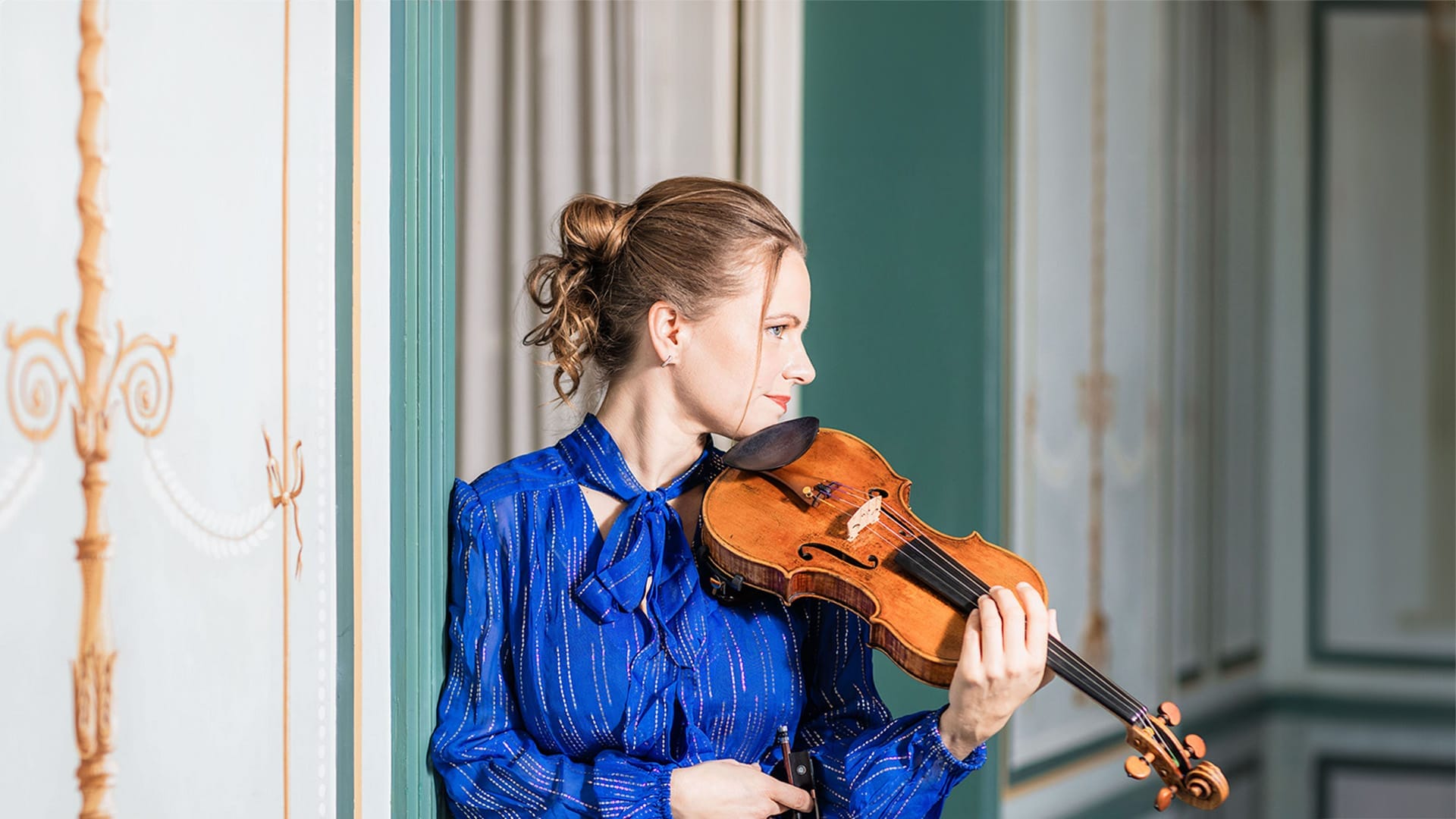 Violinist Julia Fischer, a woman with light blonde hair pulled back in a loose bun, wearing a blue blouse, holding her violin up to her shoulder, contemplative expression.