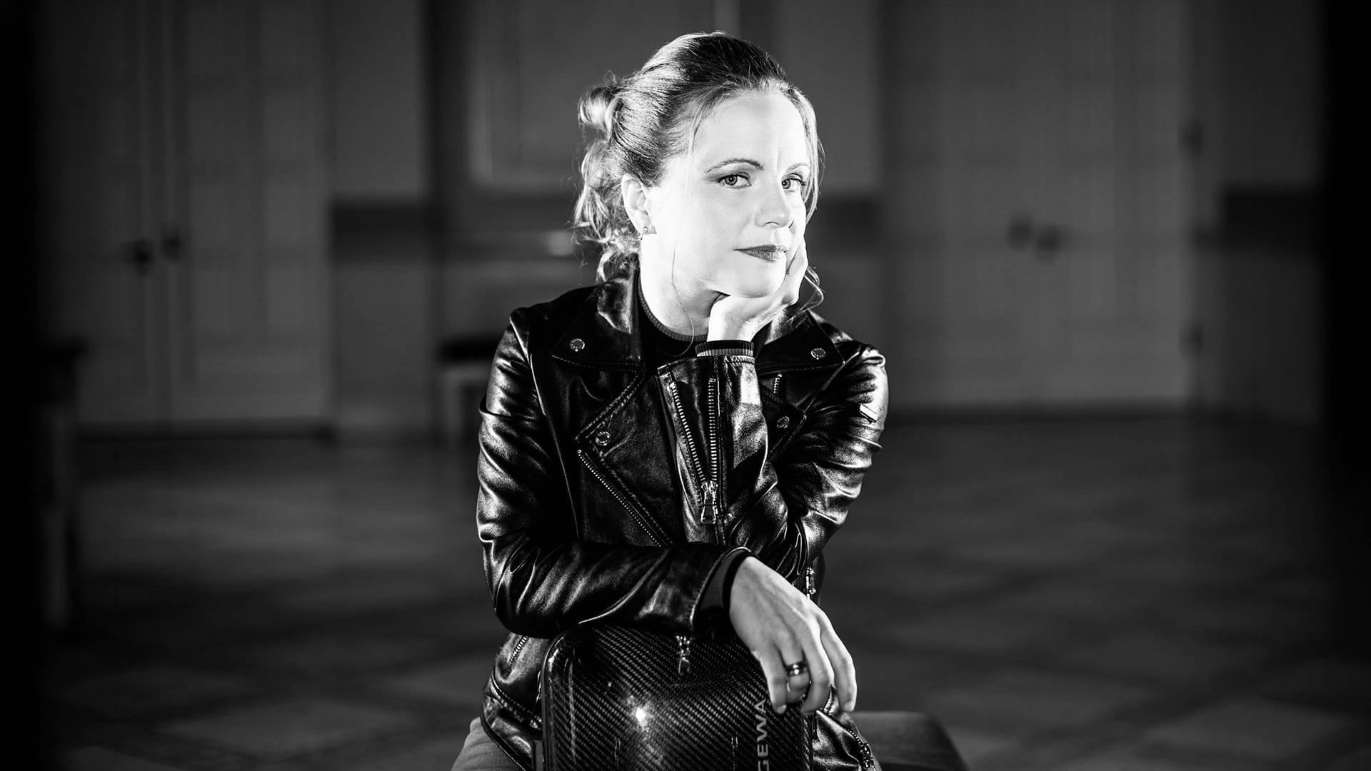 Black and white portrait of Julia Fischer, wearing a leather jacket, smiling and resting her chin on her hand.
