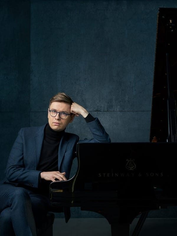 Pianist Víkingur Ólafsson, a man with short light blond hair and round glasses, wearing a dark navy suit jacket over a black turtleneck, seated beside a grand piano in a dark blue/grey studio, resting one hand against his temple in a thoughtful pose, looking directly at the camera.