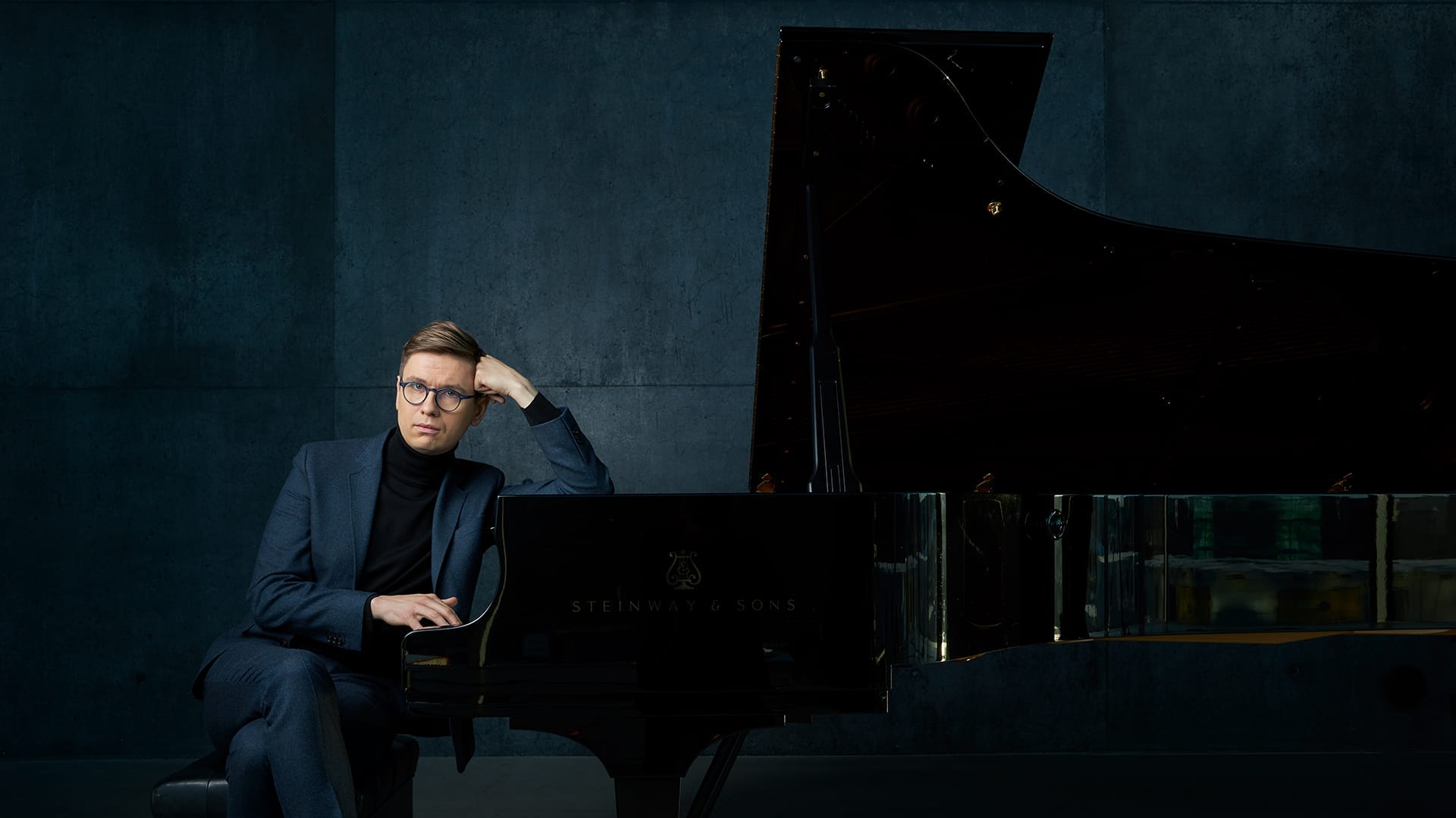 Pianist Víkingur Ólafsson, a man with short light blond hair and round glasses, wearing a dark navy suit jacket over a black turtleneck, seated beside a grand piano in a dark blue/grey studio, resting one hand against his temple in a thoughtful pose, looking directly at the camera.