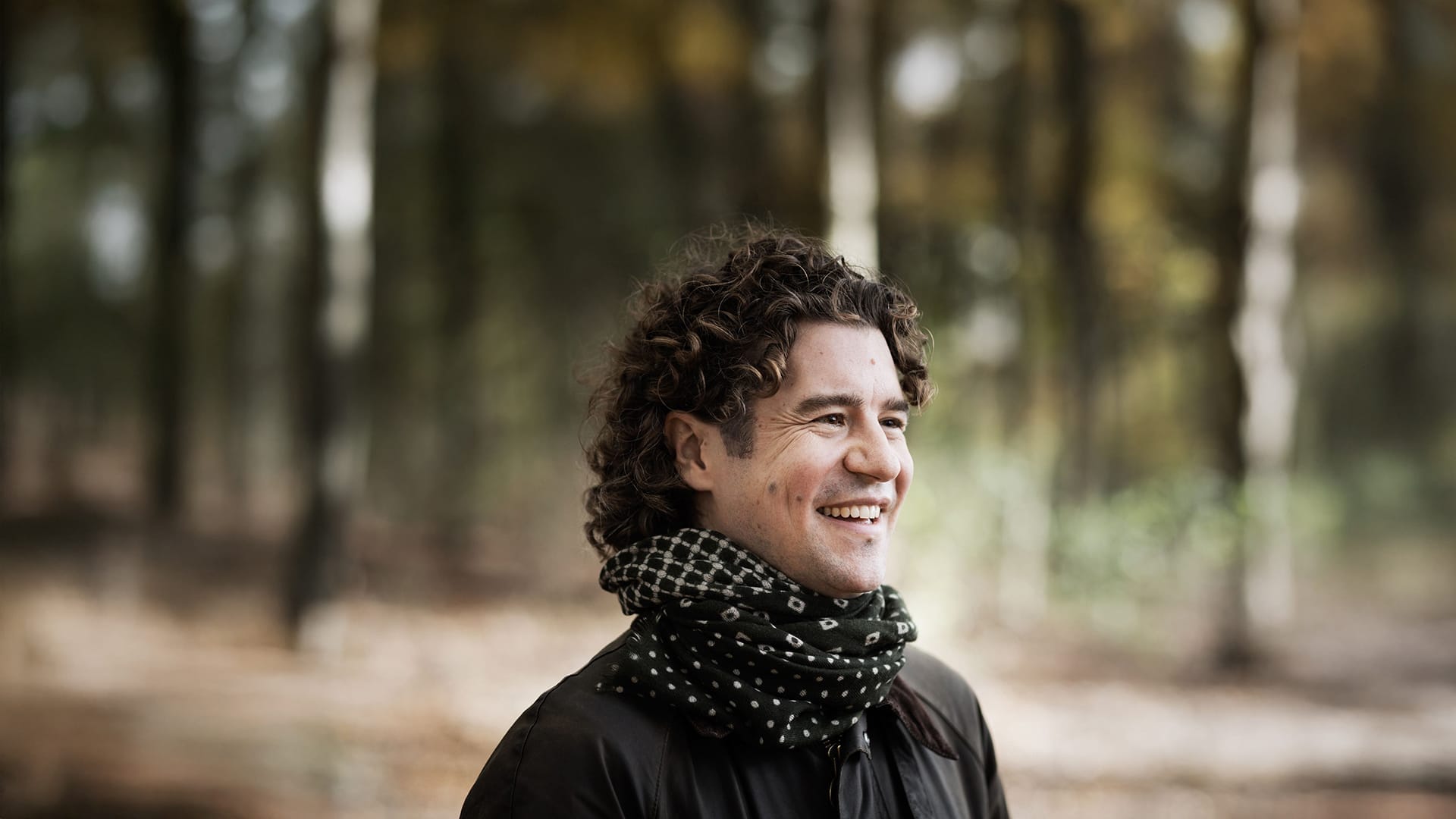 Headshot of conductor Robin Ticciati smiling outdoors, wearing a scarf and jacket.