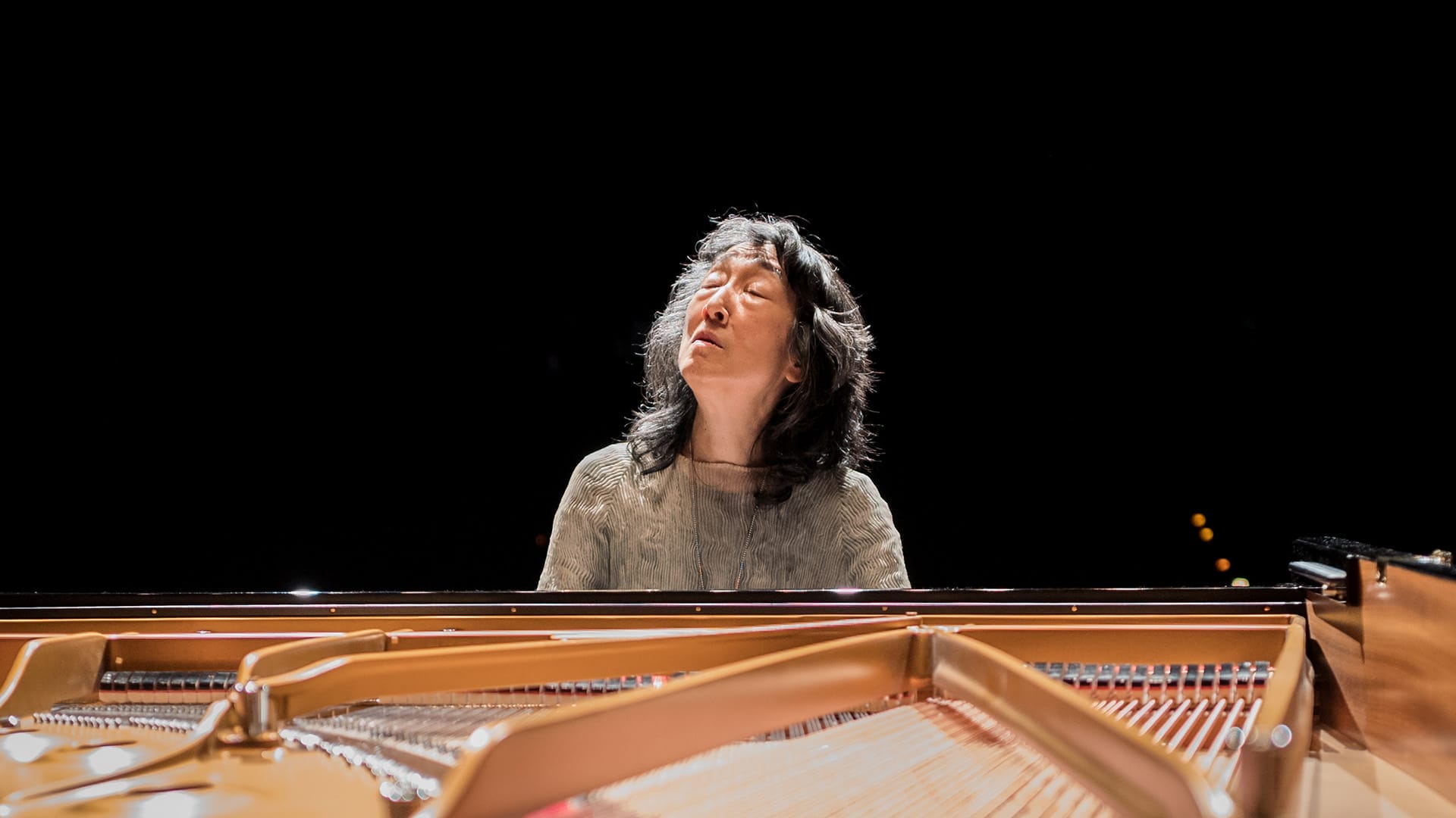 Pianist Mitsuko Uchida, a woman with shoulder-length dark grey/silver hair, wearing a grey ribbed top, seated at a grand piano with eyes closed and head tilted back in an expressive playing pose, dark background.