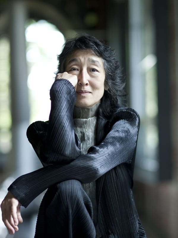 Pianist Mitsuko Uchida, a woman with dark shoulder-length hair, wearing a dark pleated jacket, seated with arms folded and chin resting on her hand, gazing ahead in a hallway with arched windows.