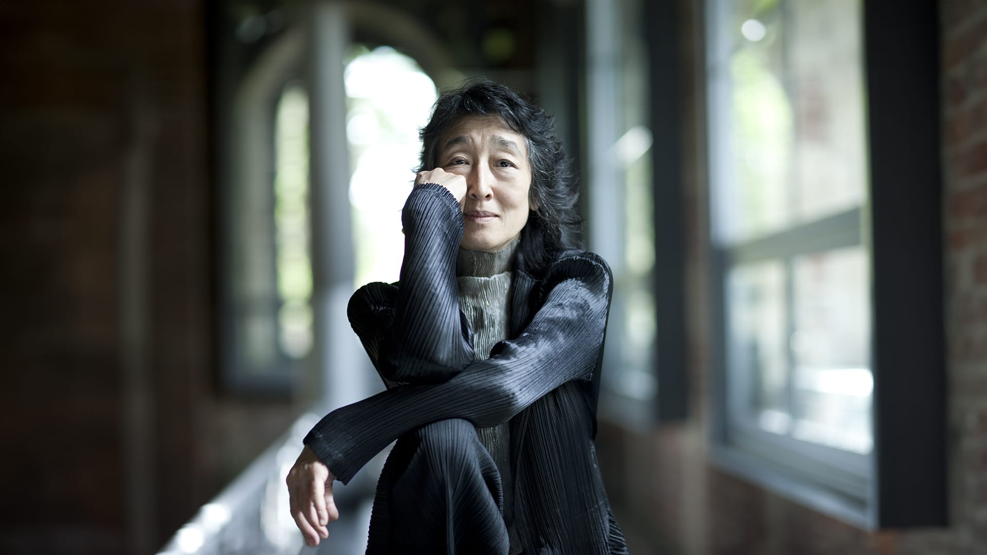 Pianist Mitsuko Uchida, a woman with dark shoulder-length hair, wearing a dark pleated jacket, seated with arms folded and chin resting on her hand, gazing ahead in a hallway with arched windows.