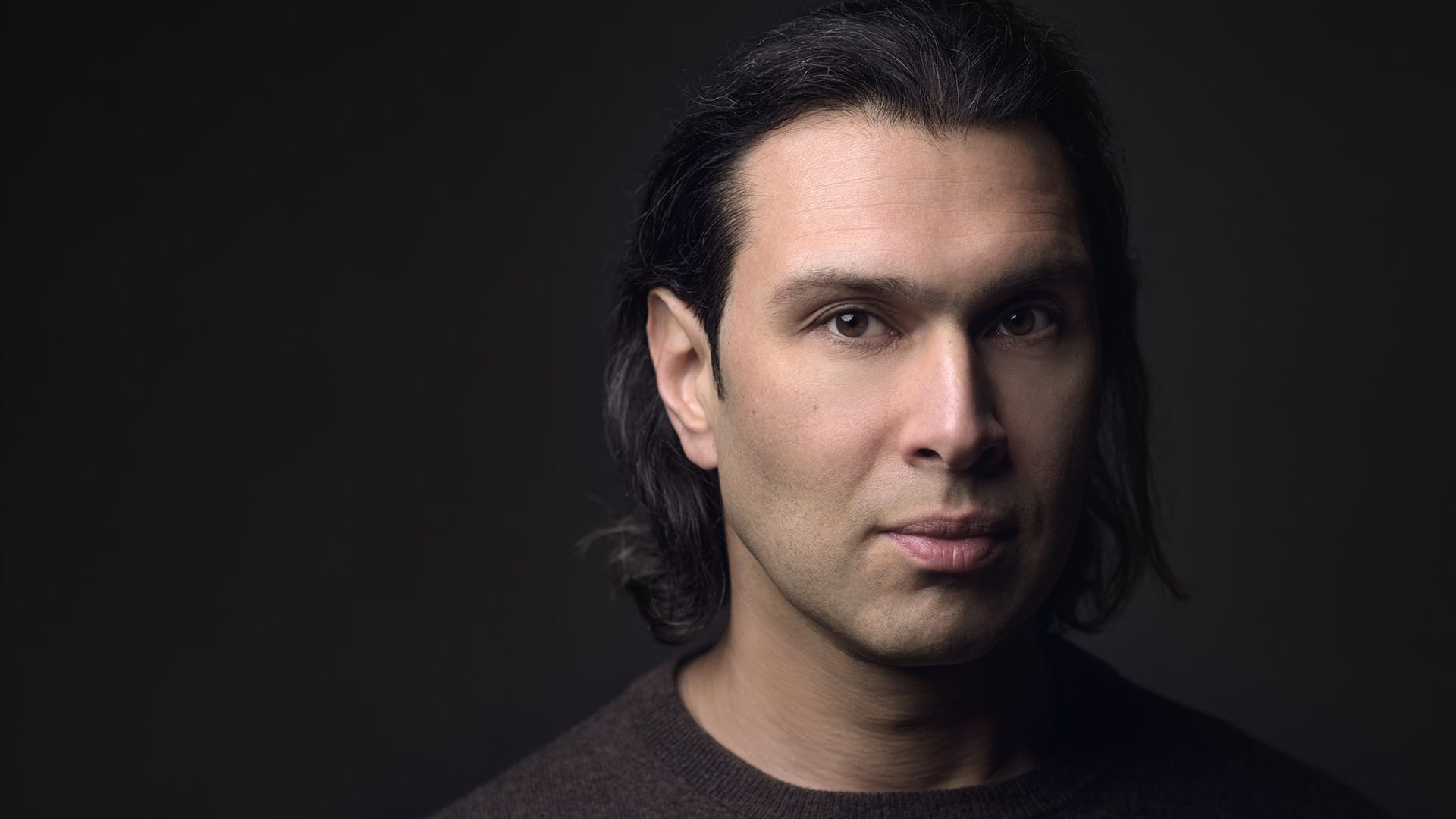 Conductor Vladimir Jurowski, a man with shoulder-length dark hair, wearing a dark top, formal close-up portrait looking directly at the camera, dark background.