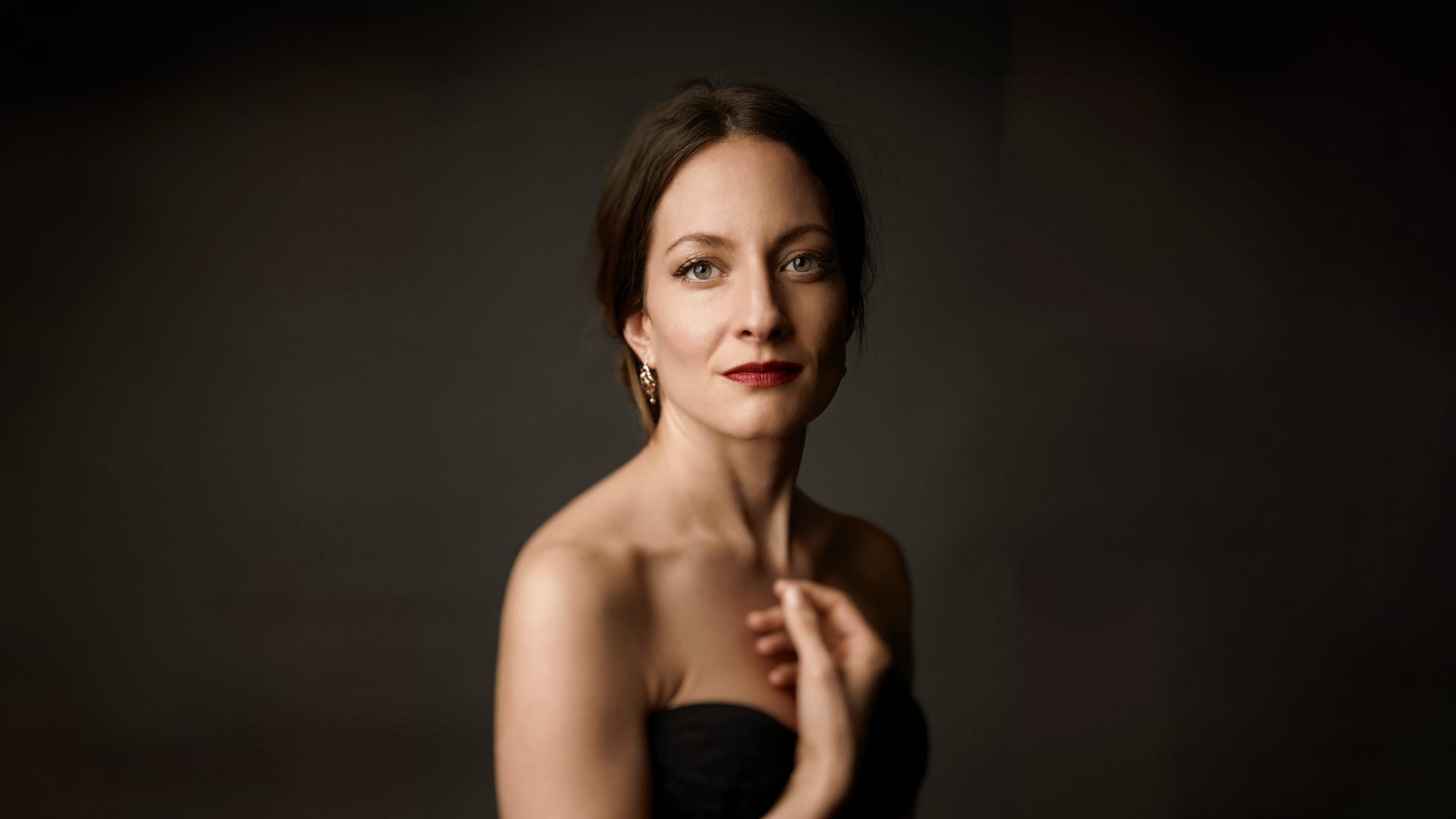 Soprano Christiane Karg, a woman with dark hair pulled back, wearing a black strapless top and red lipstick, looking gently toward the camera with one hand raised to her chest against a dark grey background.