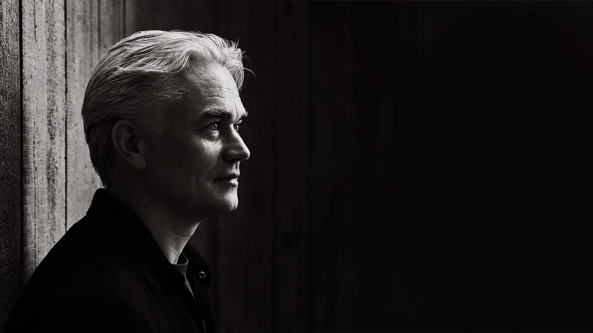 Conductor Edward Gardner, a man with light/white swept hair, conducting with a baton raised high and mouth open expressively, black and white photograph taken from orchestra level.