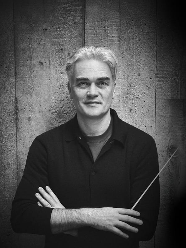 Black and white portrait of conductor Ed Gardner, a white man with short silver hair, wearing a black shirt with arms folded and holding a conductor's baton, looking at the camera against a wooden planked wall.
