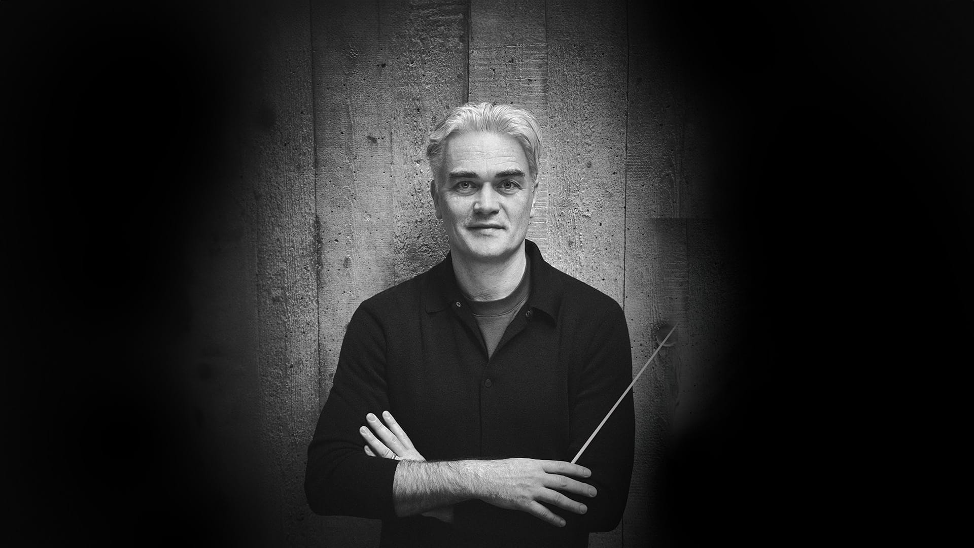 Black and white portrait of conductor Ed Gardner, a white man with short silver hair, wearing a black shirt with arms folded and holding a conductor's baton, looking at the camera against a wooden planked wall.