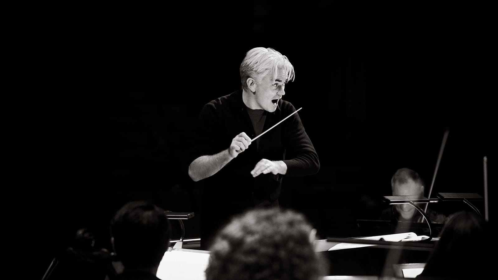 Black and white portrait of conductor Ed Gardner, a man with short silver hair, wearing a black jumper with sleeves rolled up, conducting the orchestra.