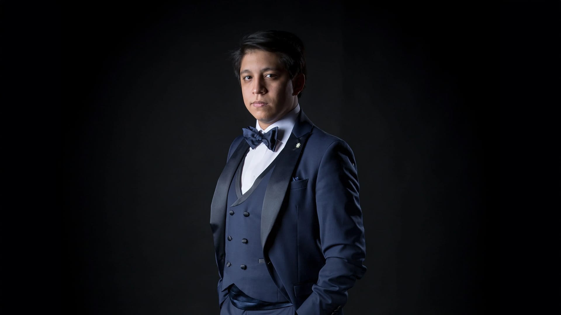 Tenor Iván Ayón-Rivas, a man with short dark hair, wearing an elegant navy three-piece suit with bow tie, standing in a formal portrait pose against a dark background.