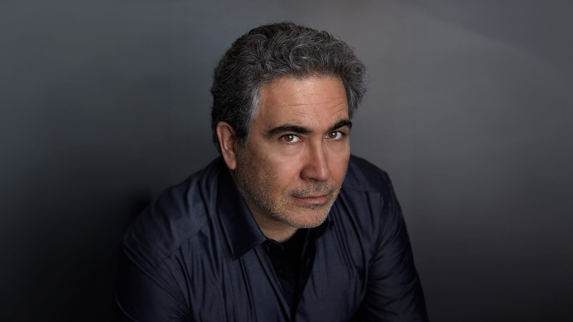 Conductor Carlo Rizzi, a man with short curly dark grey/salt-and-pepper hair and stubble beard, wearing a dark navy shirt, photographed from above looking up at the camera with an intense expression, grey background.