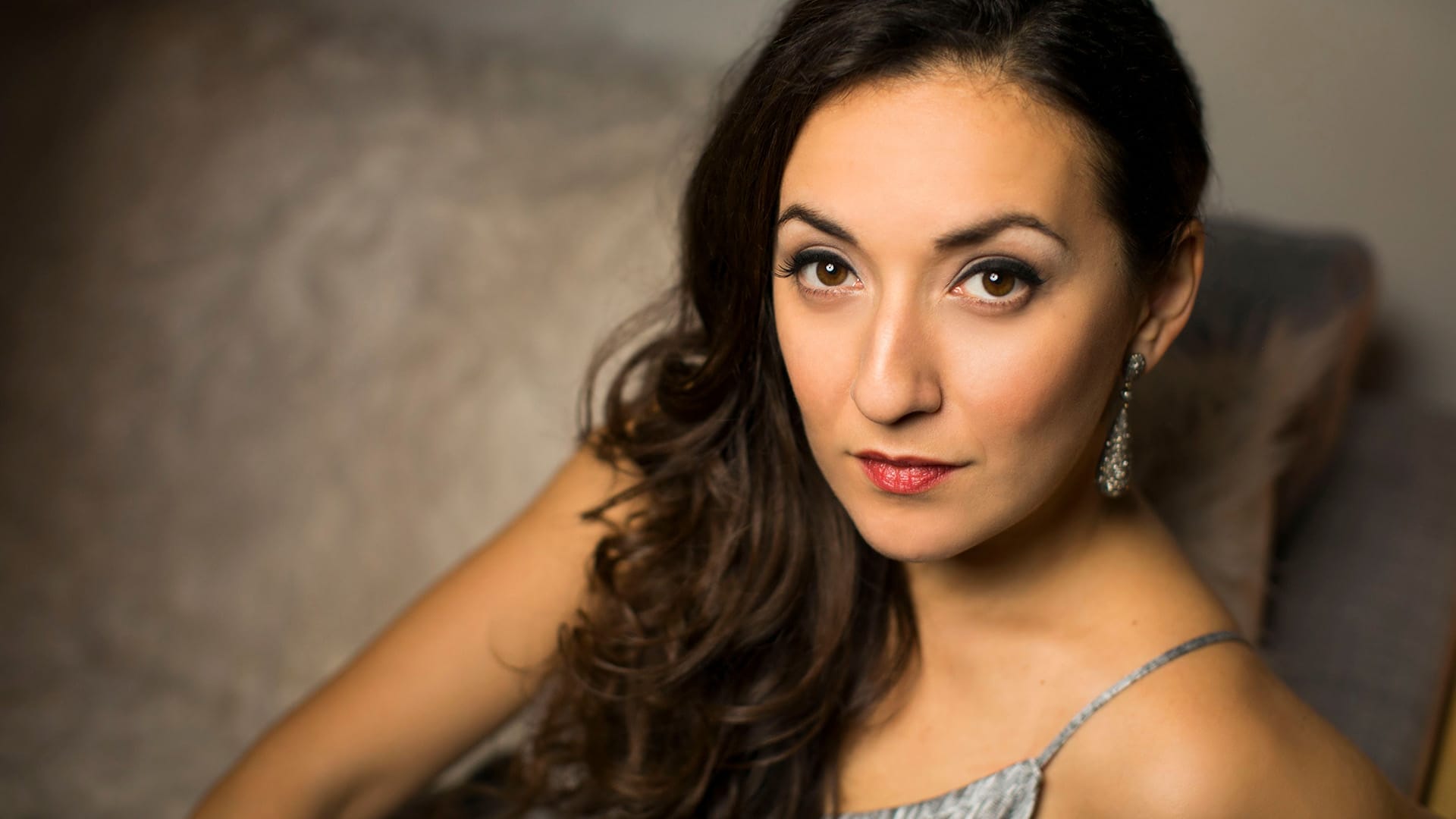 Soprano Rosa Feola, a woman with long dark wavy hair, wearing a silver/grey strappy dress with statement earrings, reclining in a chair with a direct, glamorous expression.