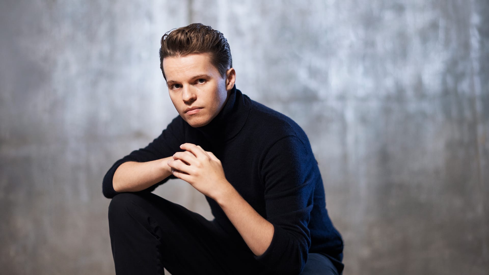Adam Hickox sits with one knee raised, hands clasped together, looking directly at the camera against a textured gray background.