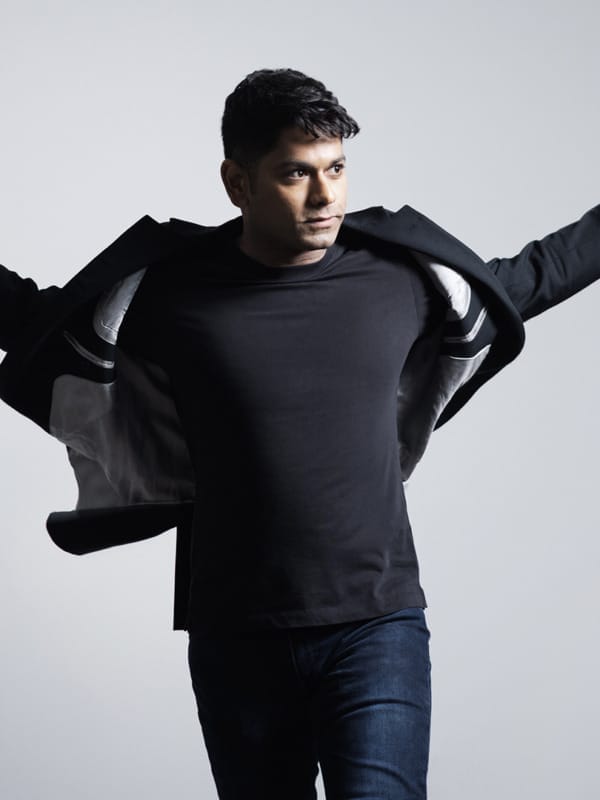 Conductor Alpesh Chauhan stands against a light gray background with his arms outstretched, looking to his right as the jacket flares slightly around him.