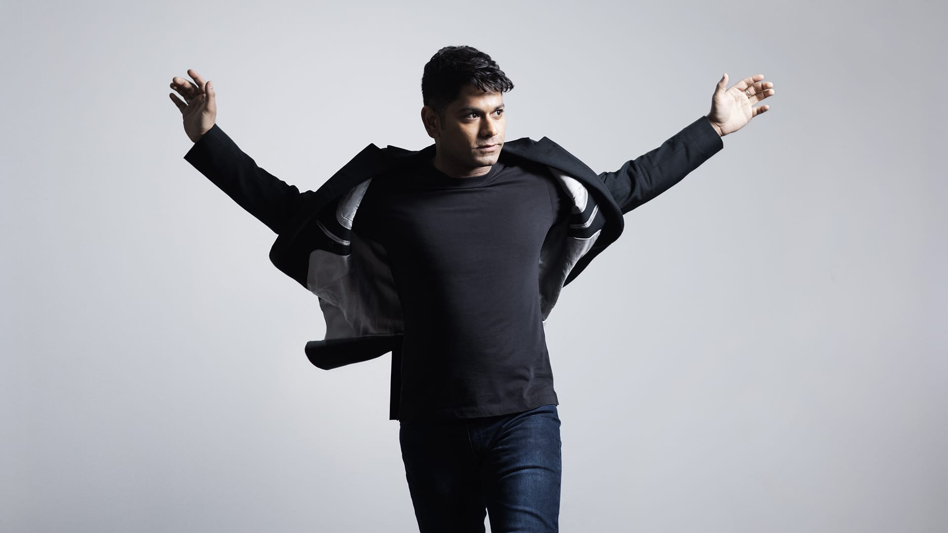 Conductor Alpesh Chauhan stands against a light gray background with his arms outstretched, looking to his right as the jacket flares slightly around him.