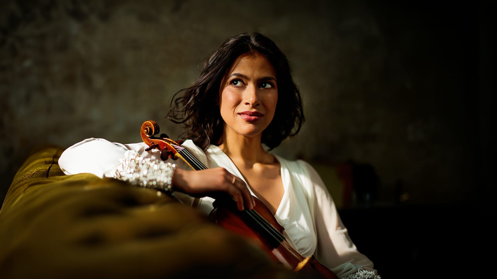 Portrait of Elena Urioste sat on a green sofa facing the camera side-on and holding her violin.