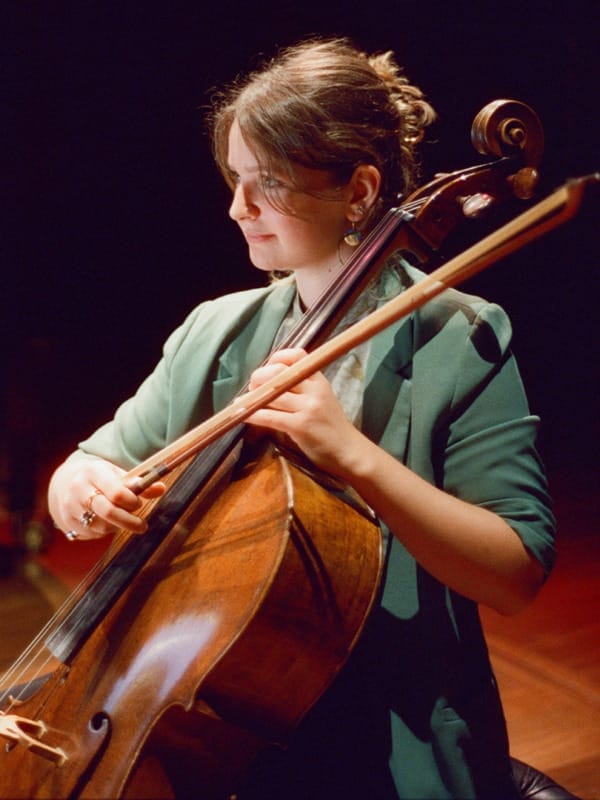 Laura van der Heijden plays her cello under warm stage lighting, wearing a green blazer and gazing down at the instrument in focused concentration as she plays.