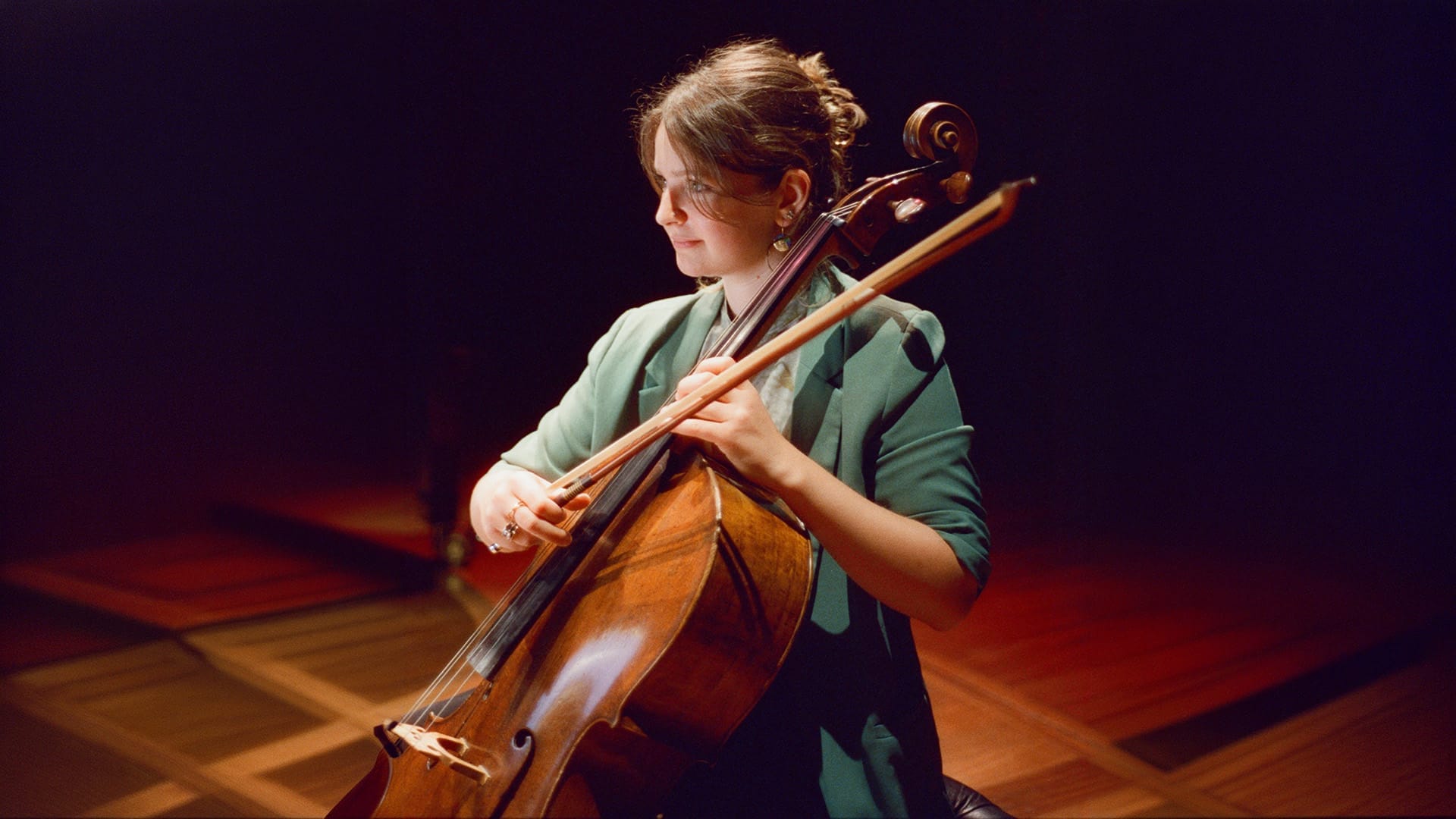 Laura van der Heijden plays her cello under warm stage lighting, wearing a green blazer and gazing down at the instrument in focused concentration as she plays.
