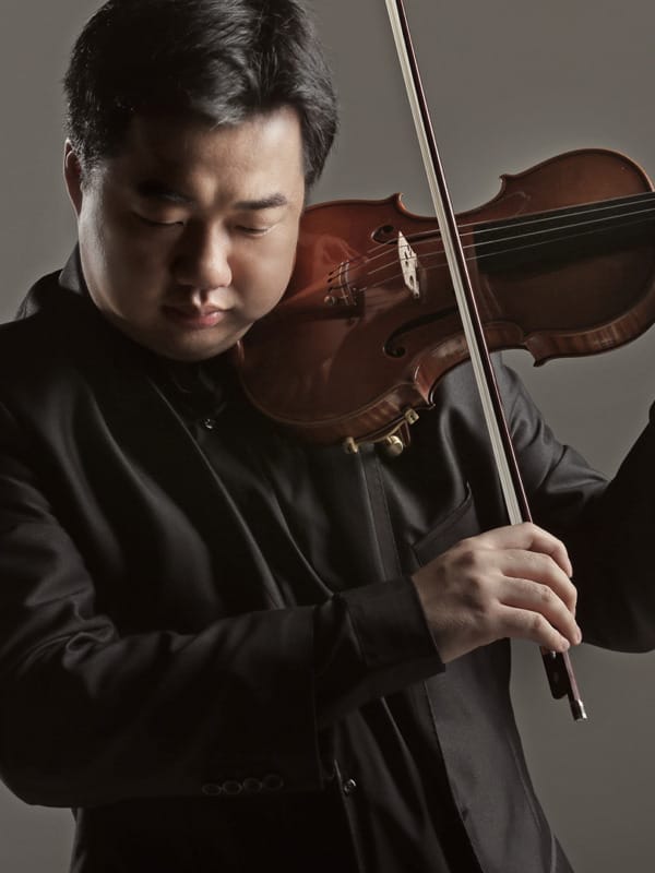 Ning Feng is captured wearing a black suit playing his violin against a plain gray background, while looking down in concentration.