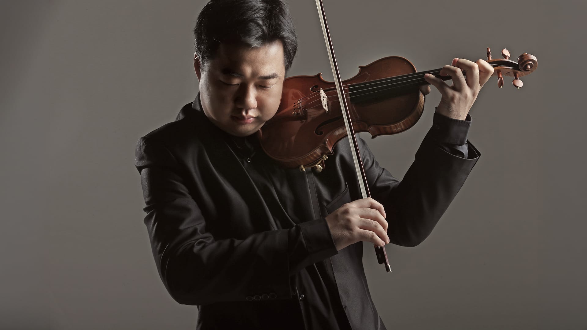 Ning Feng is captured wearing a black suit playing his violin against a plain gray background, while looking down in concentration.