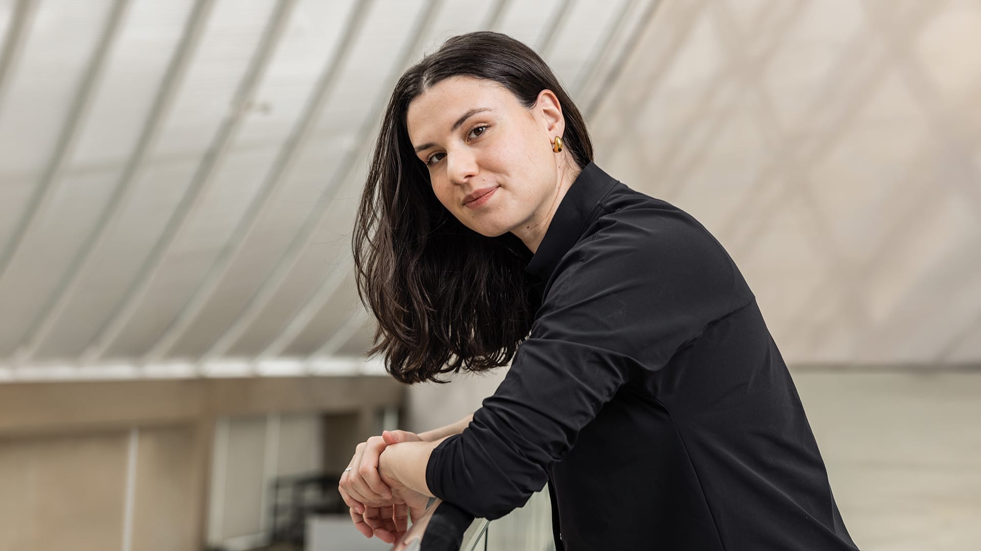 Fellow Conductor Shira Samuels-Shragg wears a black shirt, leaning forward with her hands clasped, looking at the camera with a slight smile in an indoor architectural setting.