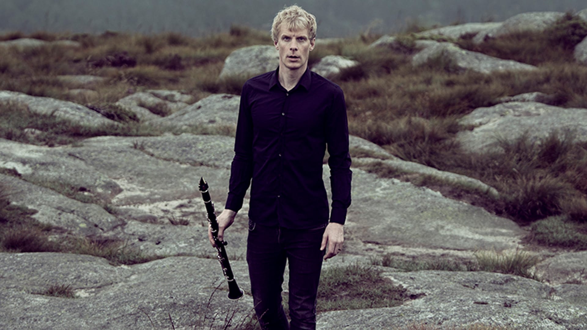 Clarinettist Martin Fröst, a man with short blond hair, wearing all black, standing on a rocky outdoor landscape holding a clarinet at his side, looking directly at the camera with a serious expression, grey overcast sky and rocky terrain behind him.