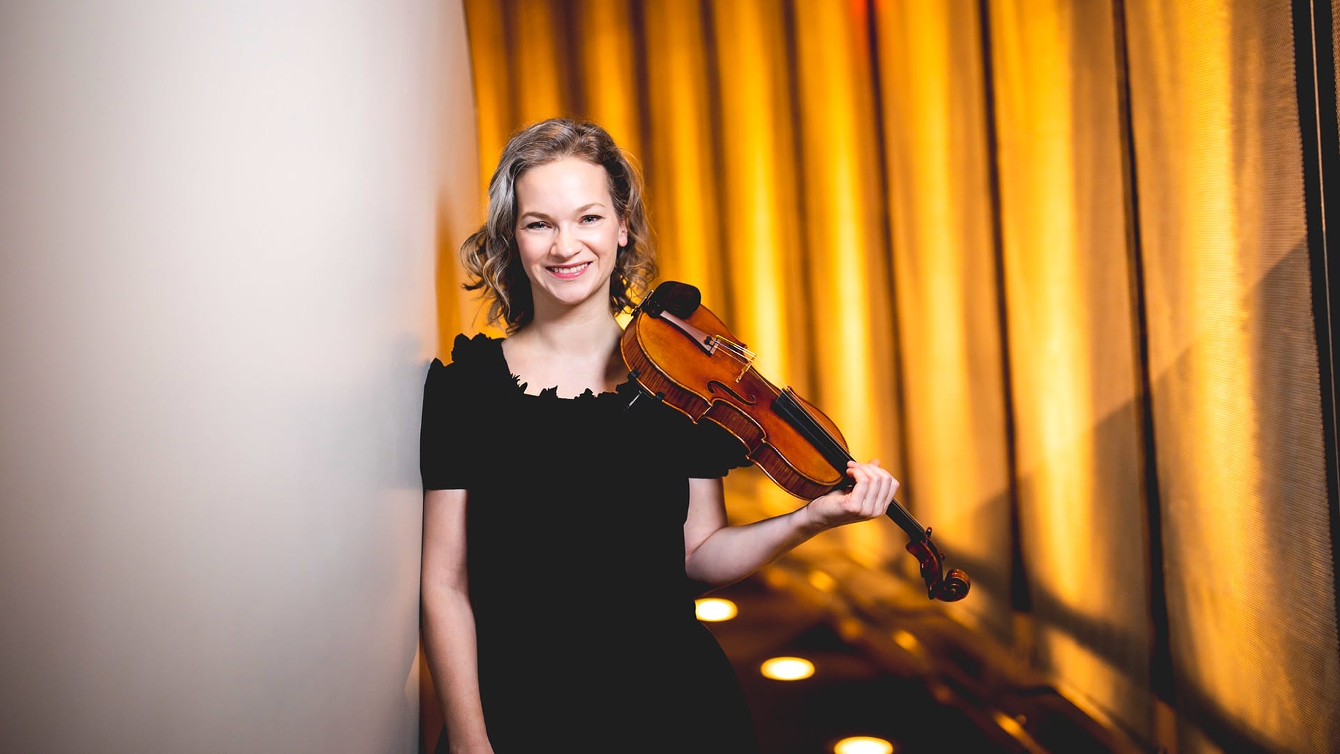 Violinist Hilary Hahn, a woman with short blonde wavy hair, wearing a black dress with a ruffle neckline, holding a violin at her shoulder and smiling, against a golden curtain backdrop.