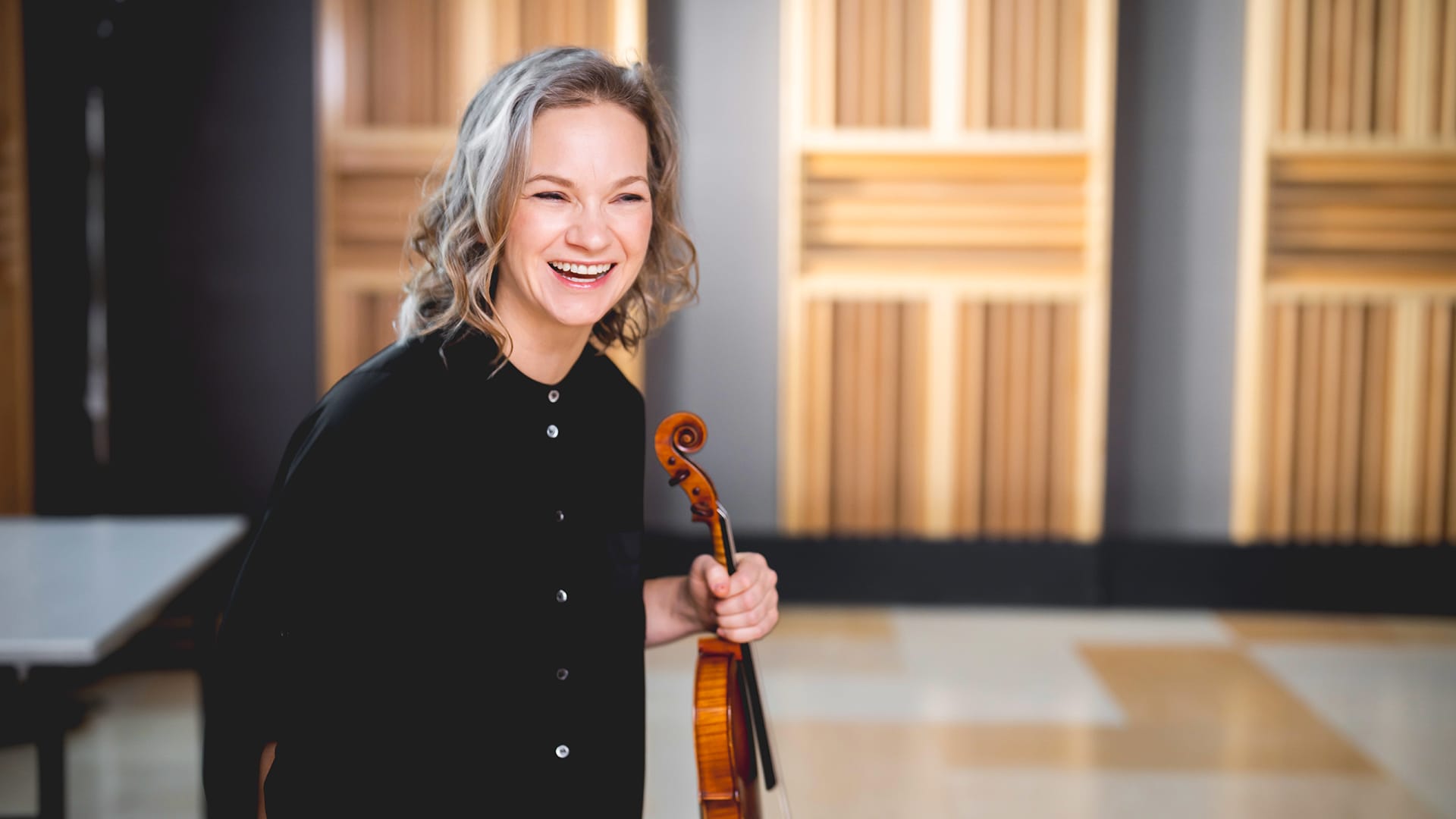 Backstage shot of Hilary Hahn holding her violin up to her shoulder and smiling to the camera.