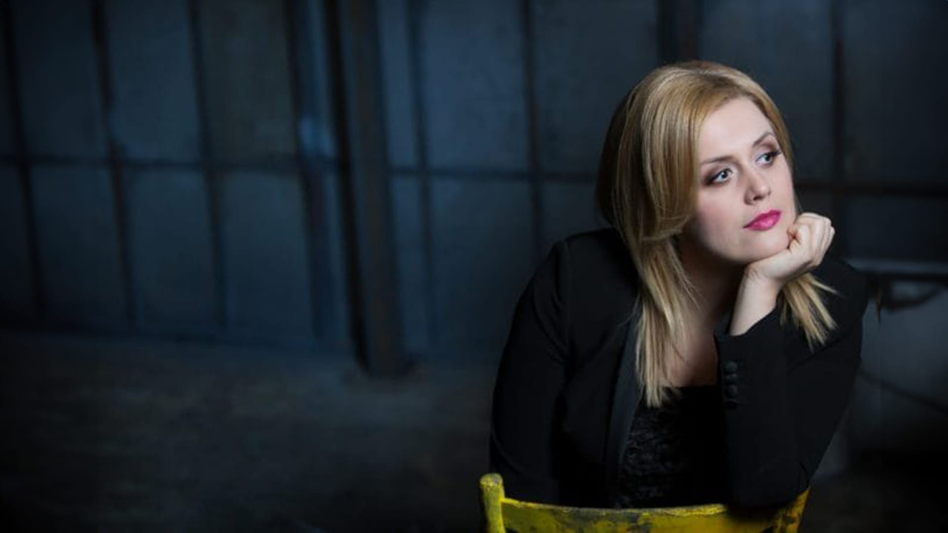 Soprano Iwona Sobotka, a woman with long straight blonde hair, wearing a black jacket, seated on a yellow chair with chin resting on hand, looking to the side thoughtfully, moody dark background.