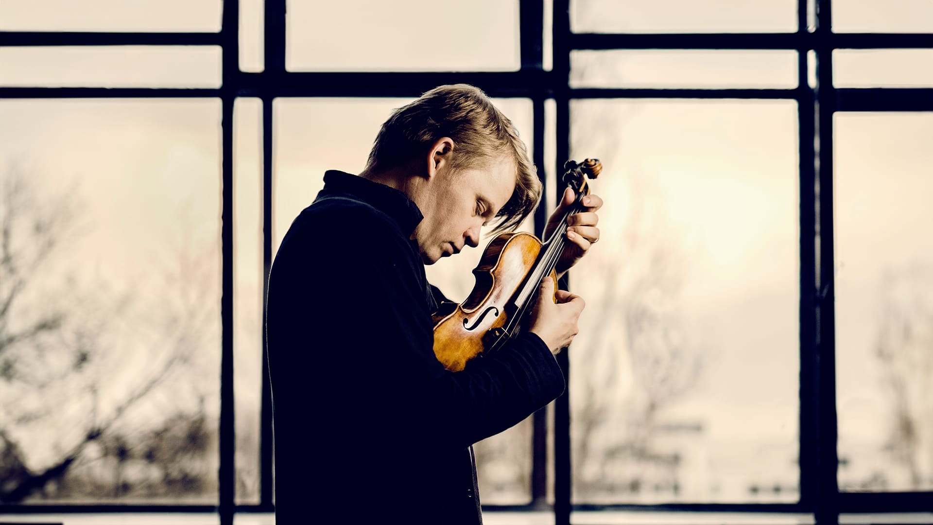Violinist Pekka Kuusisto, a man with short blonde hair wearing a dark jacket, standing in profile and holding his violin, gazing downward, against a large window with bare trees visible outside.