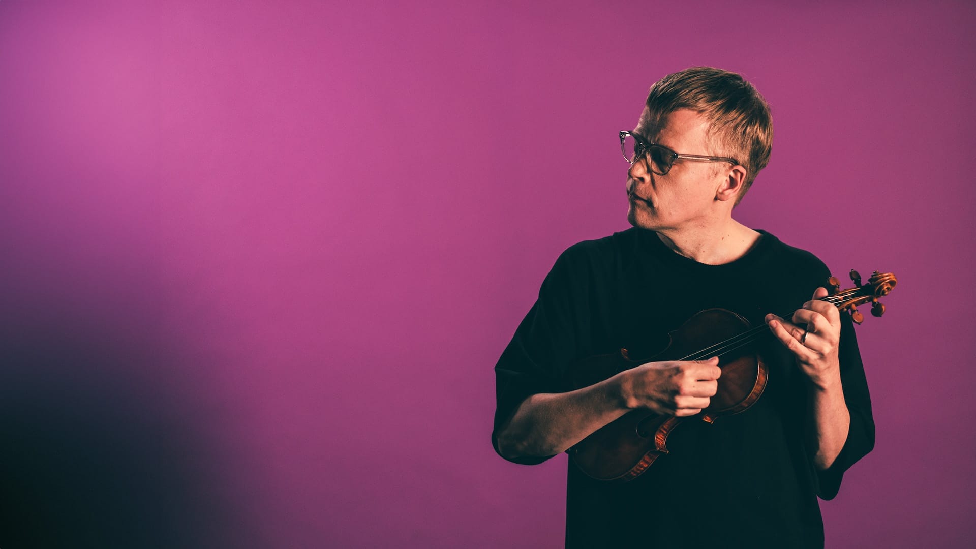 Violinist Pekka Kuusisto, a man with short blond hair and glasses, wearing a black t-shirt, holding a violin at his side and looking to the side, against a purple/magenta background.