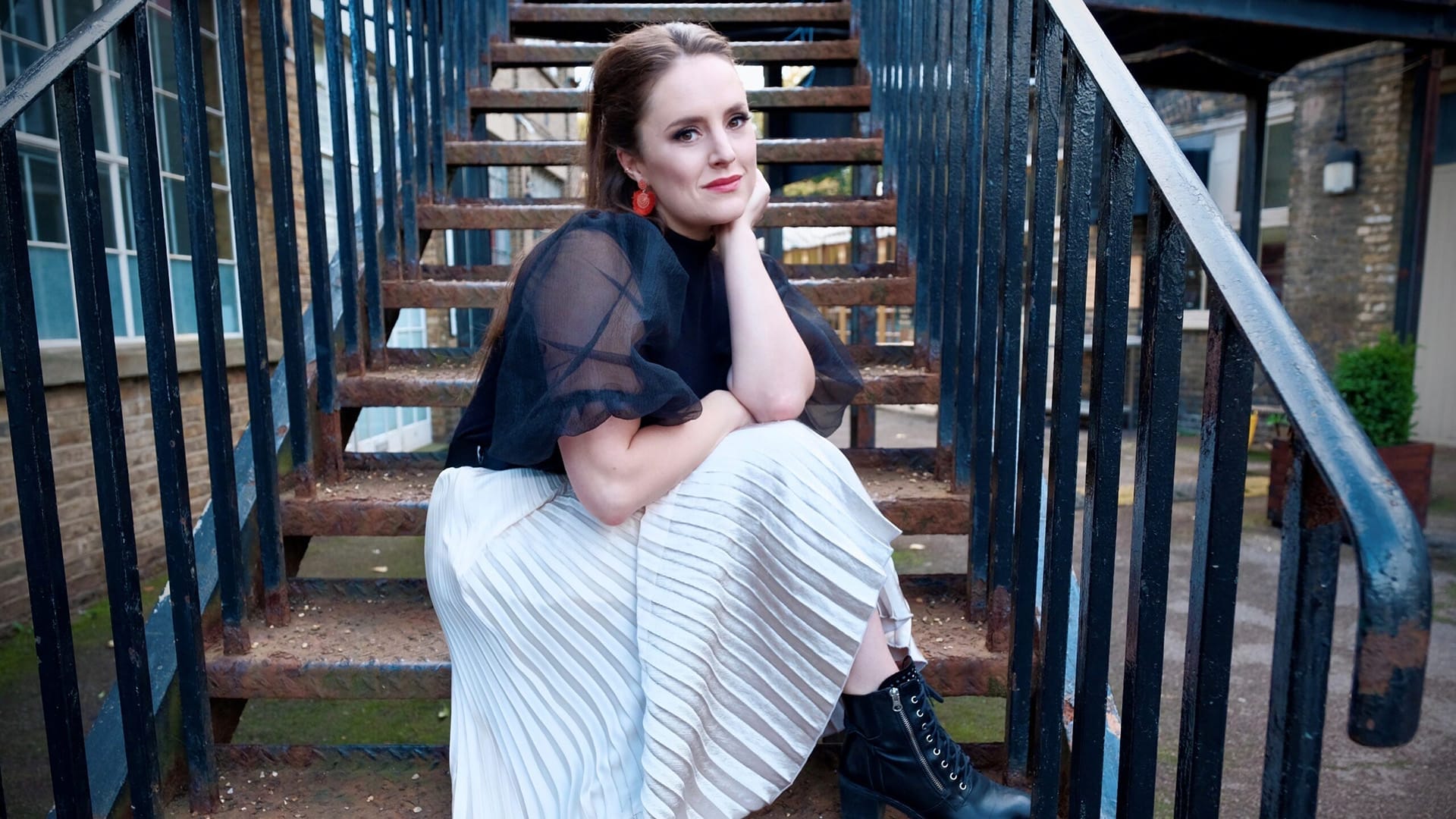 Soprano Louise Alder, a woman with dark brown hair pulled back, wearing a black sheer top and white pleated skirt with black ankle boots, seated on metal outdoor stairs, resting chin on hand, gazing thoughtfully to the side.