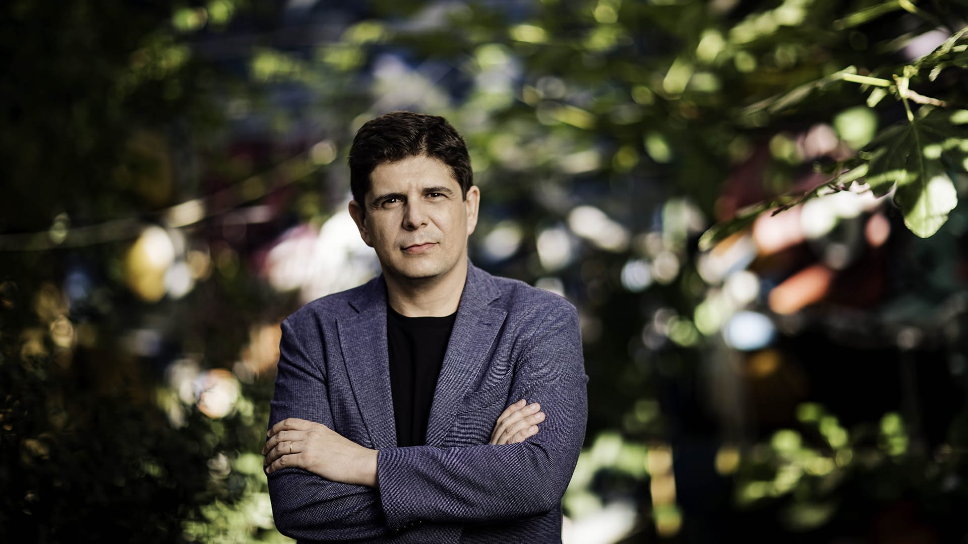 Pianist Javier Perianes, a man with short dark hair, wearing a blue/grey blazer with arms folded, standing in front of green foliage with a serious expression.