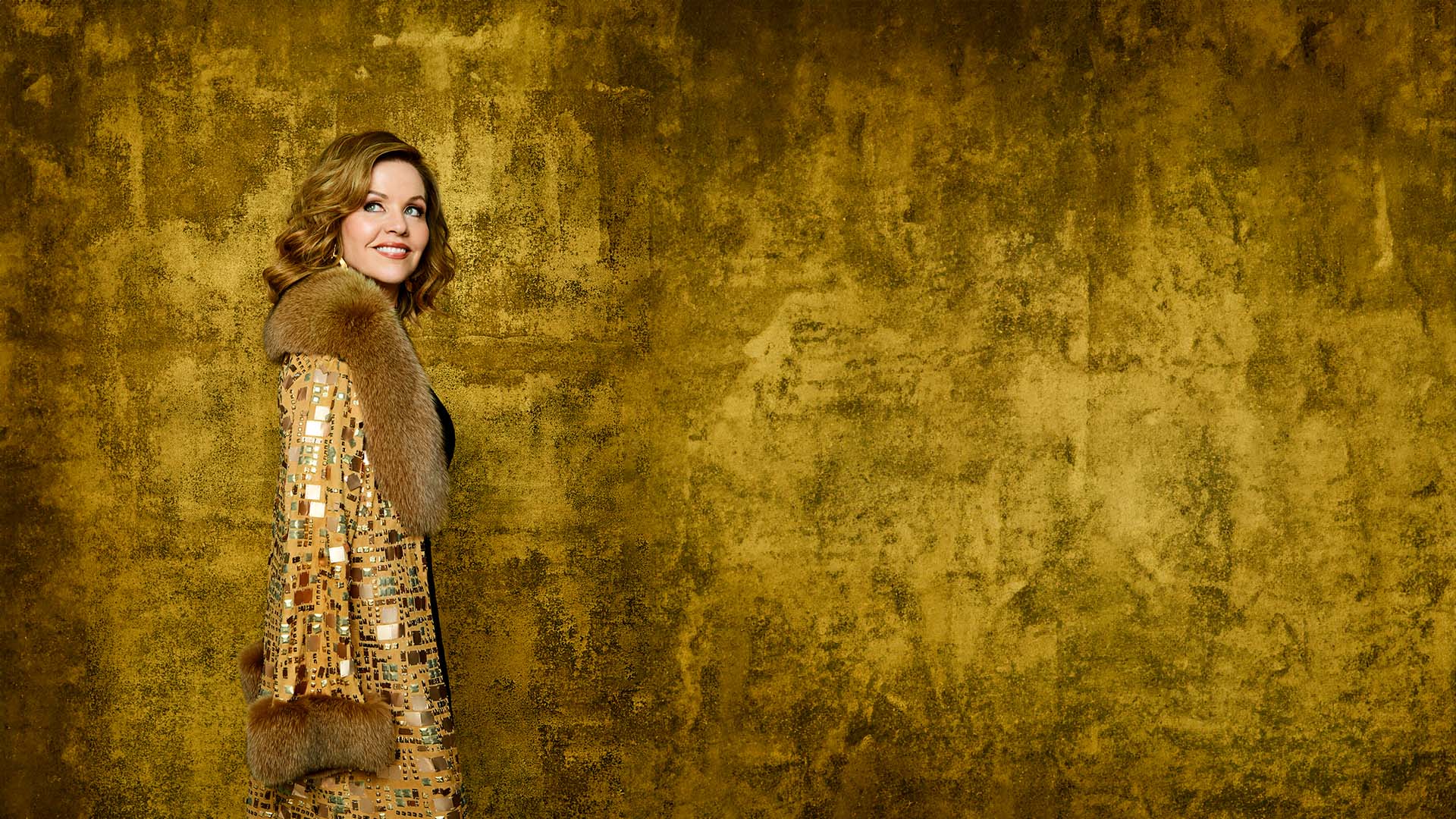 Soprano Renée Fleming, a woman with short blonde wavy hair, wearing a gold sequined coat with fur trim, standing side-on and looking back over her shoulder against a gold textured wall.