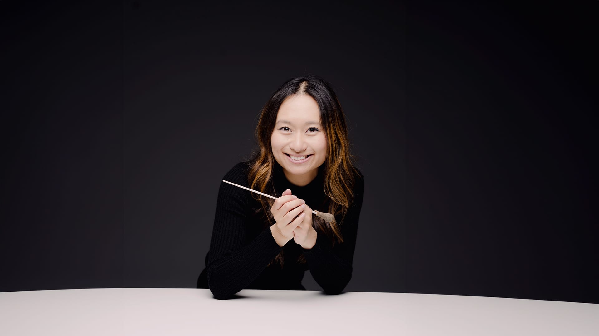 Conductor Tianyi Lu, a woman with dark hair, smiles and looks at the camera whilst leaning on a table with a conductor's baton in her hands.