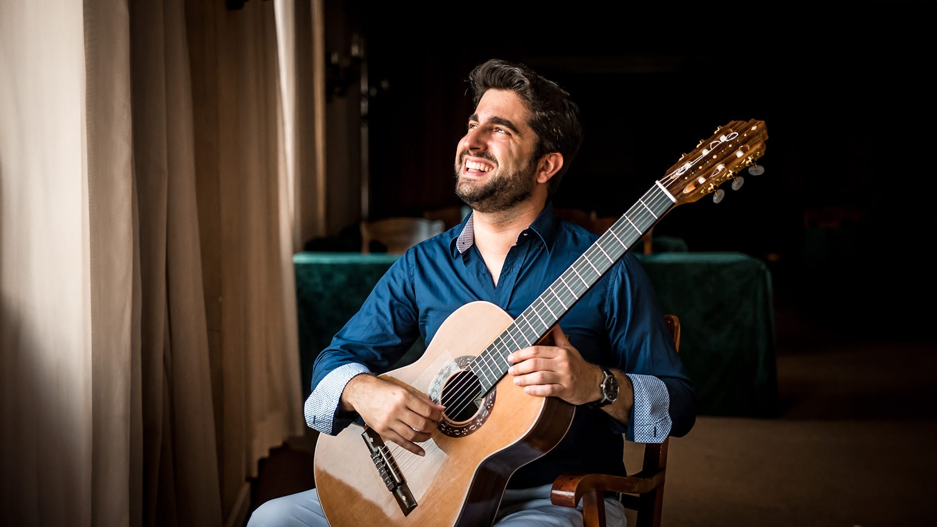 Guitarist Rafael Aguirre, a man with short dark hair and a short beard, wearing a blue shirt, seated playing a classical guitar and smiling broadly, looking upward.