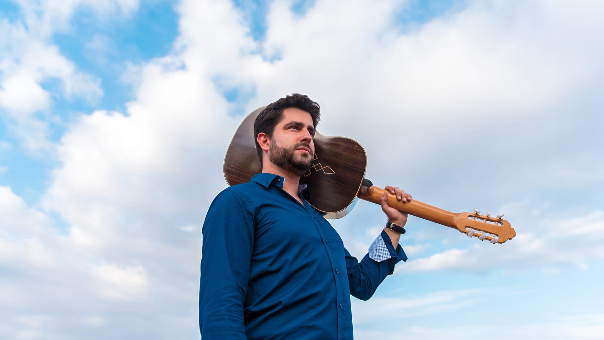 Guitarist Rafael Aguirre, a man with short dark hair and a beard, wearing a blue shirt and holding a classical guitar over his shoulder, looking into the distance against a cloudy sky.