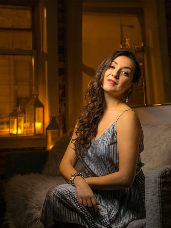 Soprano Rosa Feola, a woman with long brown, curled hair, looks at the camera whilst seated on a fur-lined sofa.