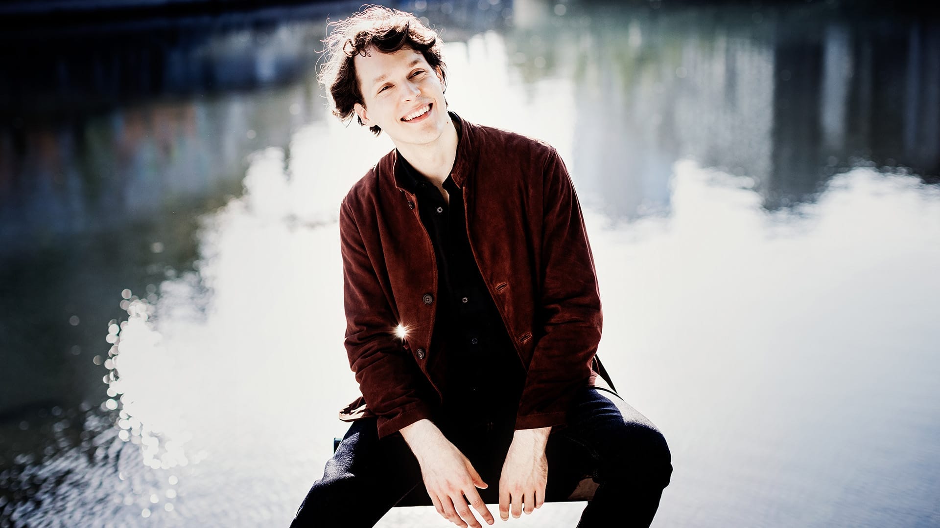 Pianist Lukas Sternath, a man with short, wavy dark hair, sits in front of a lake and smiles towards the camera.